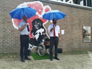Photo des deux comédiens avec des parapluies bleus devant une fresque