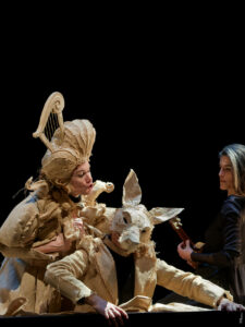 Un homme avec un masque de loup, une femme et une musicienne sont sur scène.