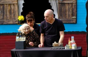 La marionnette et le comédien son derrière une table sur laquelle sont placées des maquettes.