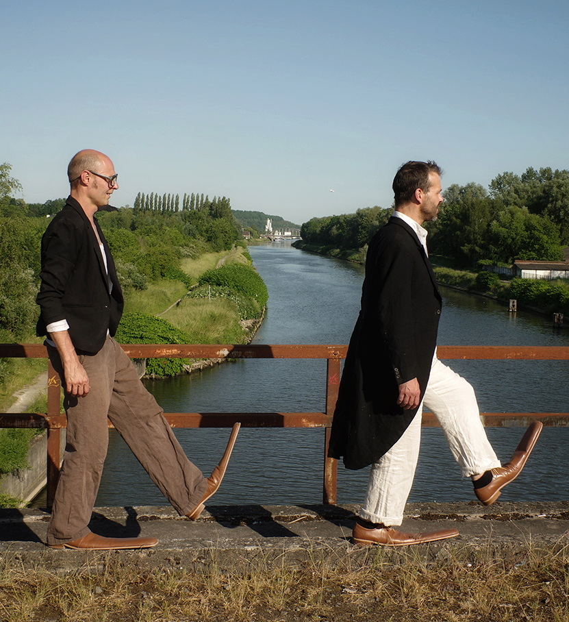 Les comédiens marchent sur un pont au-dessus d'une rivière. Ils portent des grandes chaussures.
