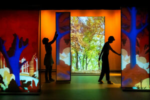 Deux personnages évoluent entre lumières et panneaux de paysages japonais colorés.