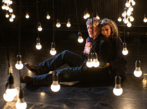 Les deux comédiens sont assis au sol et s'enlacent entre les ampoules suspendues.