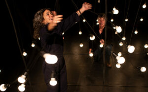 Le comédien regarde la comédienne faire bouger des ampoules suspendues.