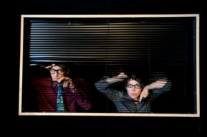 Les deux comédiens regardent le public à travers les lames d'un store.