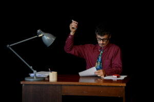 Le comédien est assis à un bureau et écrit sur une feuille.