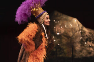 La comédienne porte un manteau en fourrure orange et une coiffe en plumes violettes.