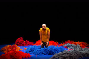 Le comédien est perplexe, agenouillé au milieu de tas de laine de couleur.