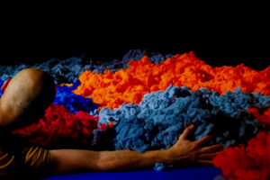 Le comédien est allongé dans de la laine de couleur qu'il entoure de ses bras.