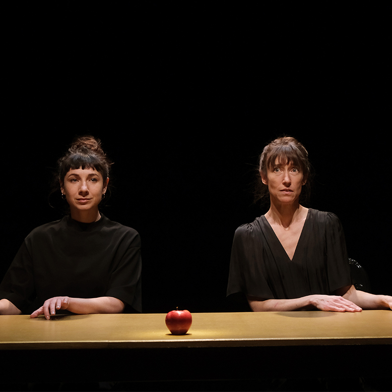 Les deux comédiennes sont assises à une table. Une pomme est posée sur la table.