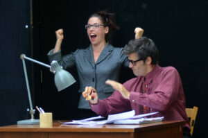 Les deux comédiens sont derrière un bureau, la comédienne fait une expression de victoire.