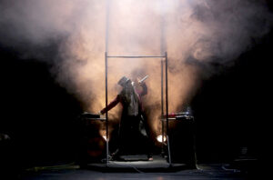 Le comédien chante entre ses deux tables de mixage avec de la fumée derrière lui.