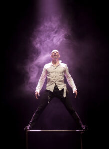 Le comédien regarde vers le ciel, il a les jambes écartée et de la fumée derrière lui.