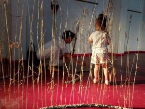 Une enfant regarde une plasticienne en train de planter un épi de blé dans de l'argile.