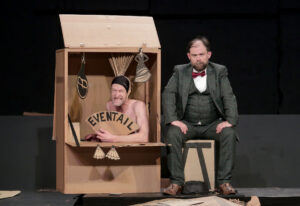 L'un des deux comédien est dans une cabane en carton. L'autre, en costume, est assis à coté.