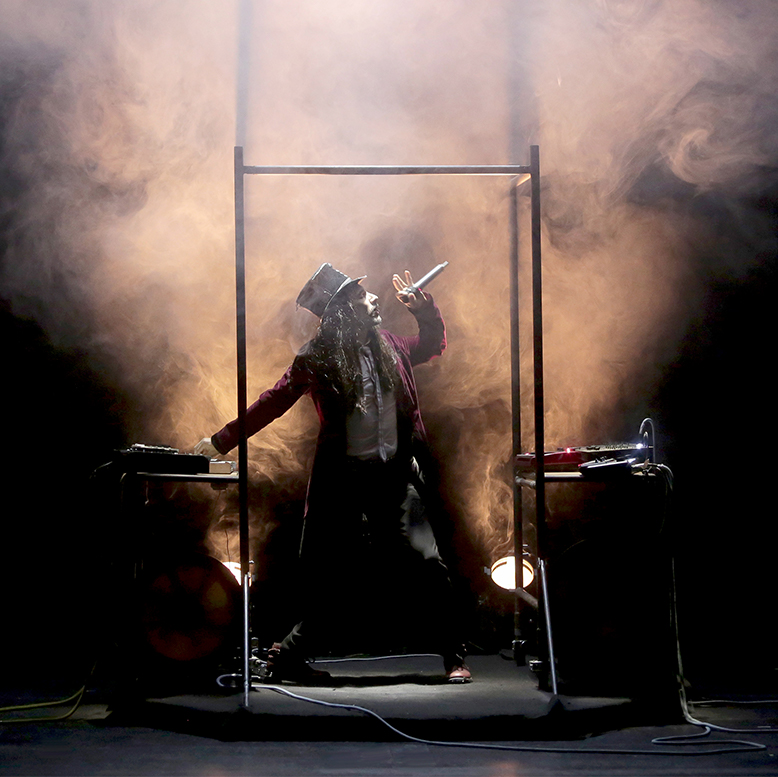 Le comédien chante entre deux claviers, il porte un chapeau et un grand manteau.