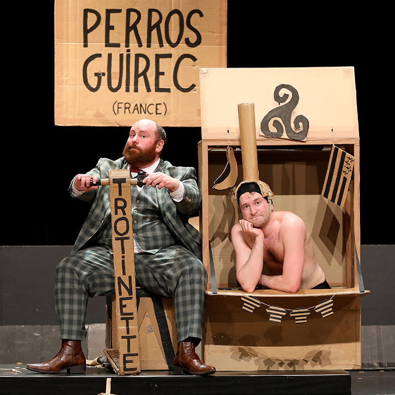 Les deux comédiens sont assis et regardent le public. Ils sont entourés de cartons.