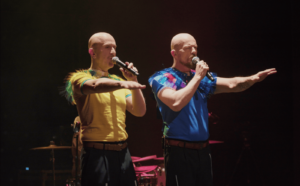 Deux musiciens chantent sur scène, en tendant leurs bras vers l'avant.