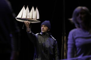 Le comédien, habillé en marin, tient une maquette de bateau dans sa main.