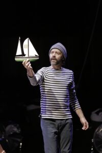 Le comédien tend une maquette de bateau vers le public.