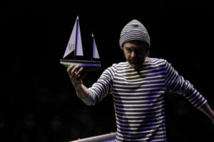 Le comédien tient une maquette de bateau dans sa main.