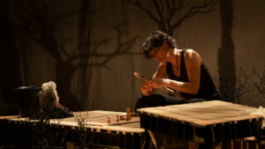 La comédienne est assise et tient un bout de bois.
