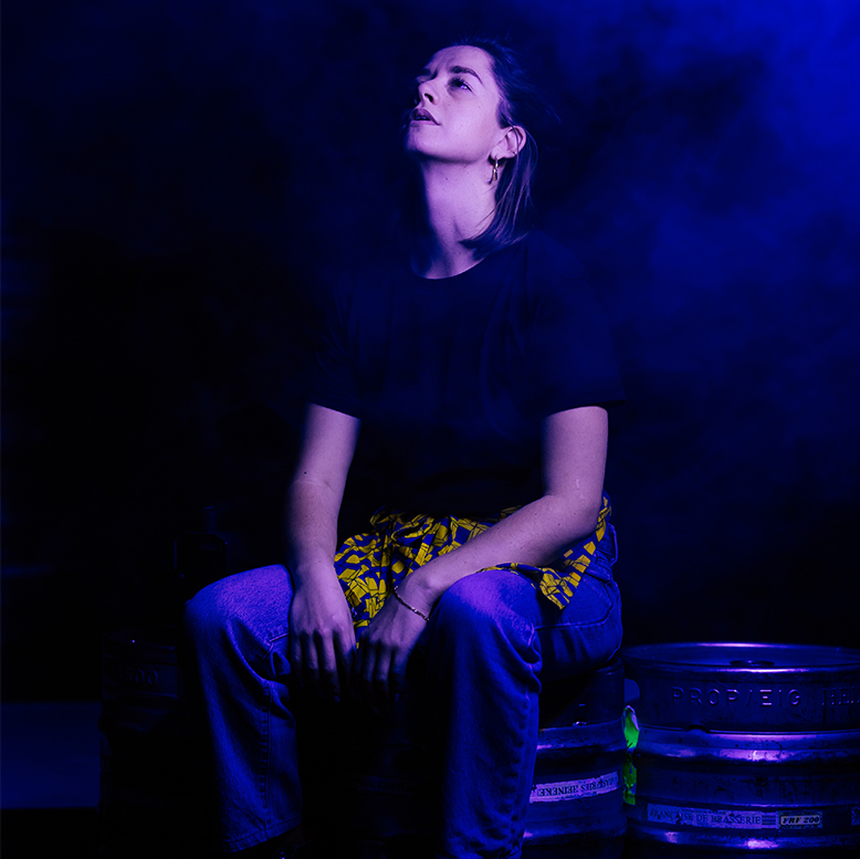 La comédienne est assise sur un fût de boisson et regarde au ciel.