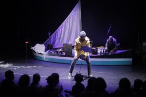 Le comédien est devant un bateau et manipule une marionnette de poisson.