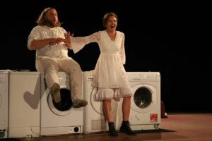 Le comédien est assis sur une machine à laver, la comédienne tend le bras vers lui.