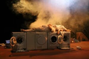Le comédien est couché sur quatre machines à laver, de la fumée derrière lui.